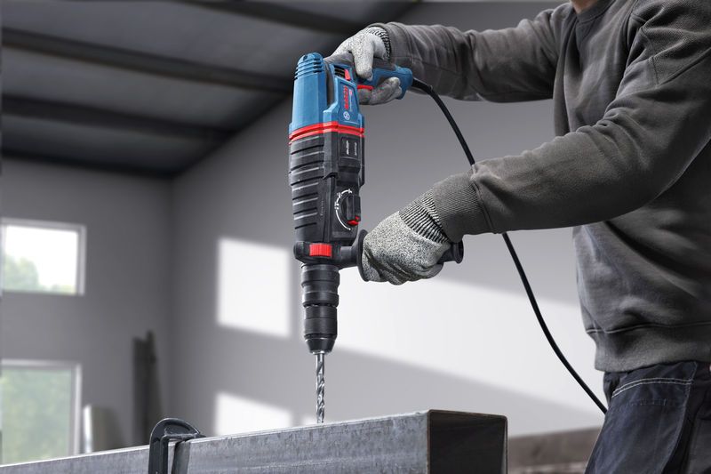 Uma pessoa que utilize equipamento de segurança perfura uma viga metálica utilizando um martelo rotativo.