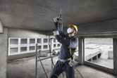 Uma pessoa que utilize equipamento de segurança perfura um teto de betão com um martelo rotativo.