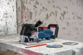 Utensílio elétrico para fresar paredes, com dois discos de corte sobressalentes e um lápis vermelho sobre uma mesa poeirenta.