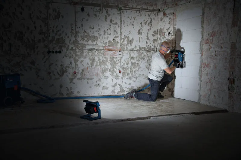Uma pessoa que usa equipamento de segurança corta canais numa parede, iluminada por uma luz de trabalho sem fios.