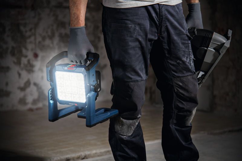 Uma pessoa que utilize equipamento de segurança segura uma luz de trabalho sem fios e uma ferramenta elétrica.