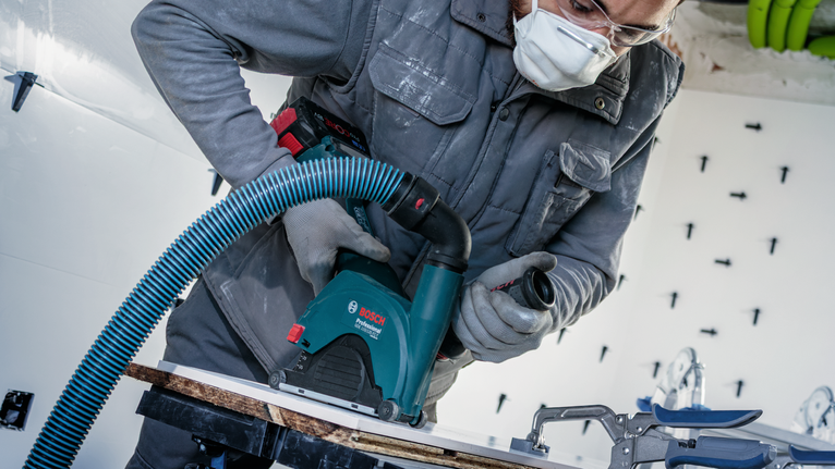 Uma pessoa que utiliza equipamento de segurança corta um painel com uma serra elétrica ligada a uma mangueira de aspirador.