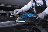 Uma pessoa que use equipamento de segurança lustra a superfície de um automóvel com uma máquina de polir sem fios.