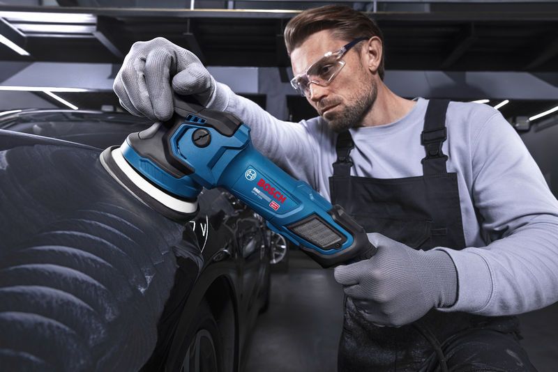 Uma pessoa que usa equipamento de segurança lustra um carro preto com uma máquina de polir elétrica.