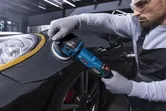 Uma pessoa que utilize equipamento de segurança utiliza uma polidora no guarda-lamas de um automóvel.