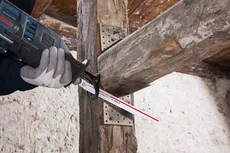 Person wearing safety equipment uses a reciprocating saw to cut a wooden beam.