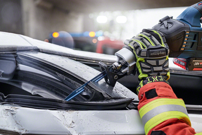 A person wearing safety equipment uses a reciprocating saw to cut through a damaged car roof.