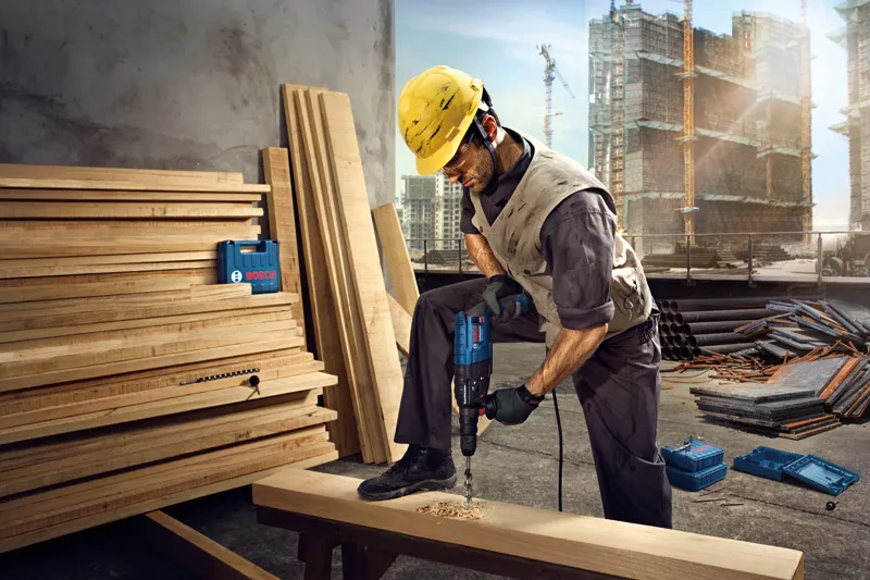 A person wearing safety equipment drills into a wooden beam at a construction site.