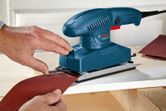 A person attaches sandpaper to an orbital sander on a wooden surface.