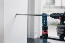 Person wearing safety equipment drills into a white wall with a power tool.