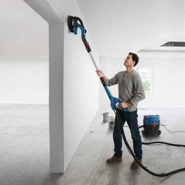 A person sands a white wall with a drywall sander connected to a dust extractor.