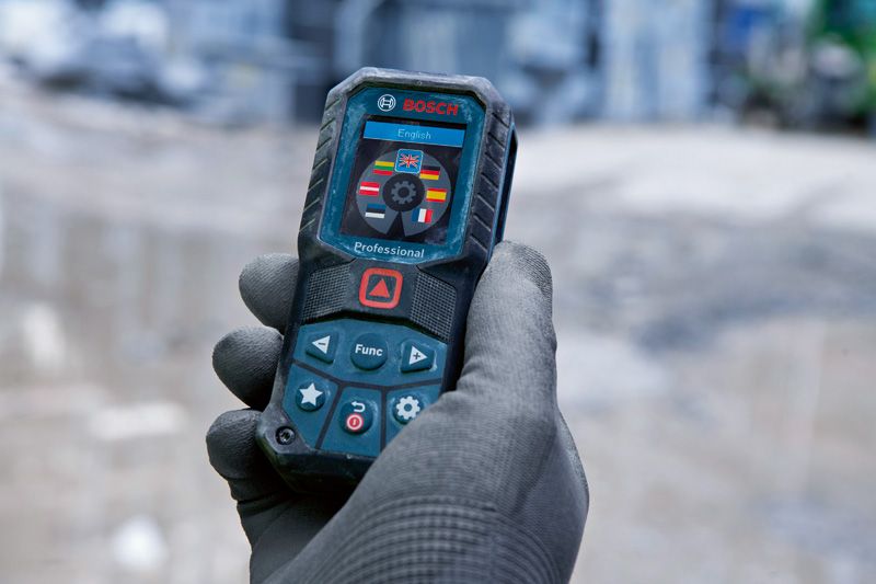 A person wearing safety equipment holds a laser measure with language selection on the screen.