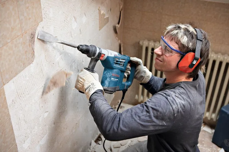 A person wearing safety equipment uses a rotary hammer to remove tiles from a wall.