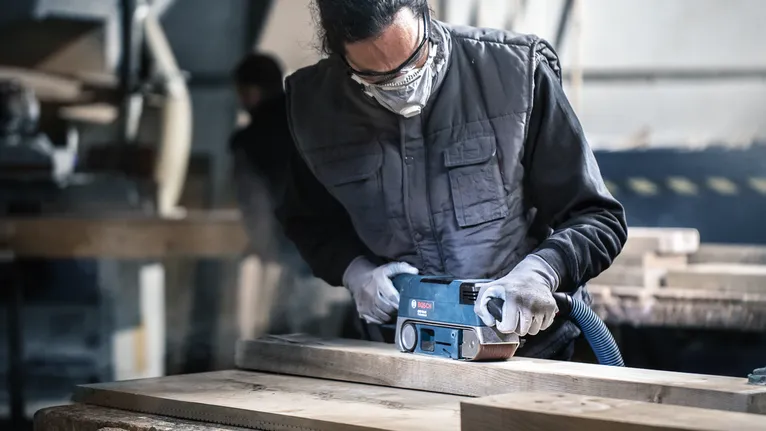 Person wearing safety equipment uses a belt sander to smooth wooden planks.