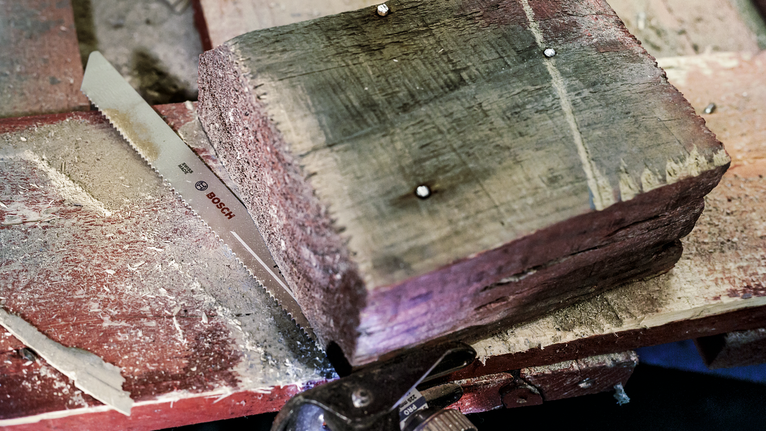Reciprocating saw blade cutting through a thick wooden beam on a workbench.