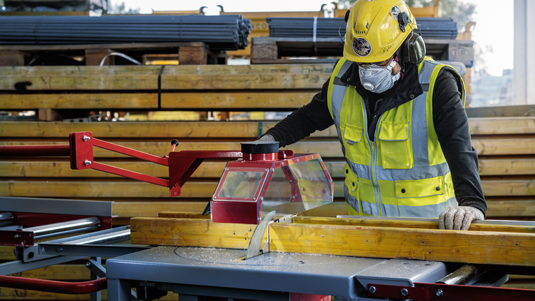 A person wearing safety equipment cuts wood with a table saw in a construction area.