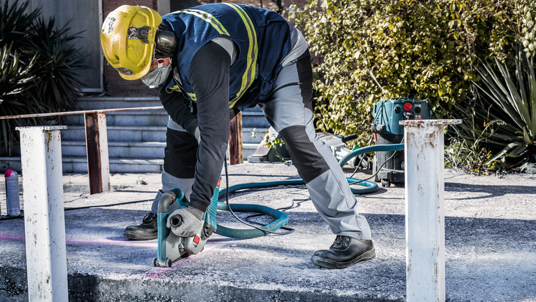 A person wearing safety equipment cuts concrete outdoors with a power saw.