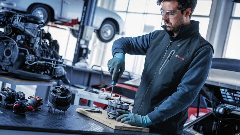 Person wearing safety equipment repairs an automotive part at a workbench.