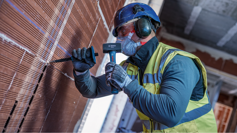 A person wearing safety equipment chisels a brick wall with a hammer.