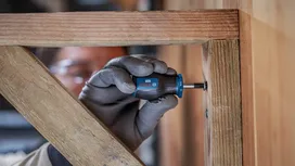 Person wearing safety equipment uses a screwdriver to fasten a screw in a wooden frame.
