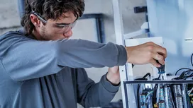 Person wearing safety equipment tightens a screw on an electrical control panel.