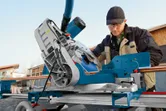 A person wearing safety equipment operates a mitre saw to cut wood on a jobsite.