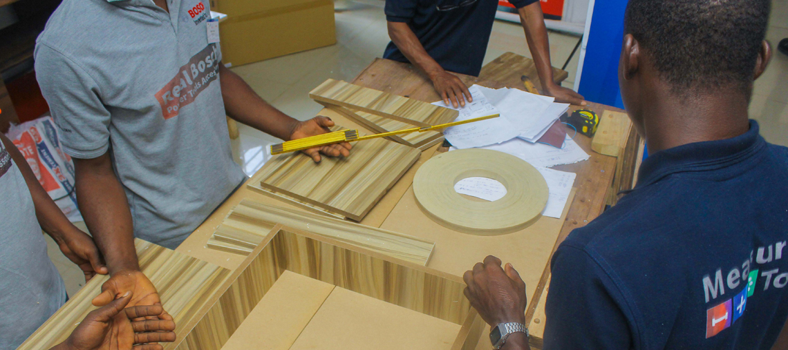 Two men work on wooden pieces with tools at a table in a workshop setting.