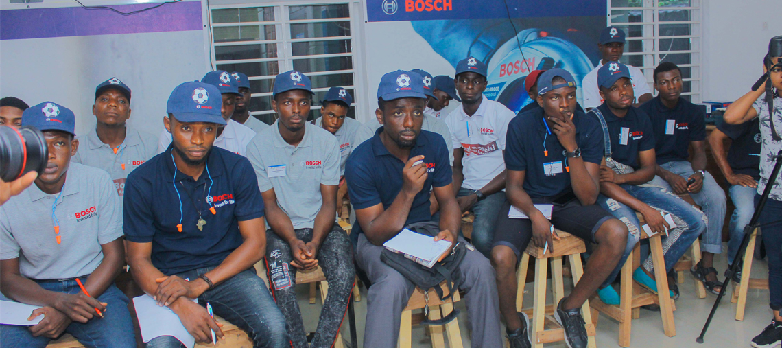 A group of men in Bosch caps and shirts sits on stools, engaged in a workshop discussion.