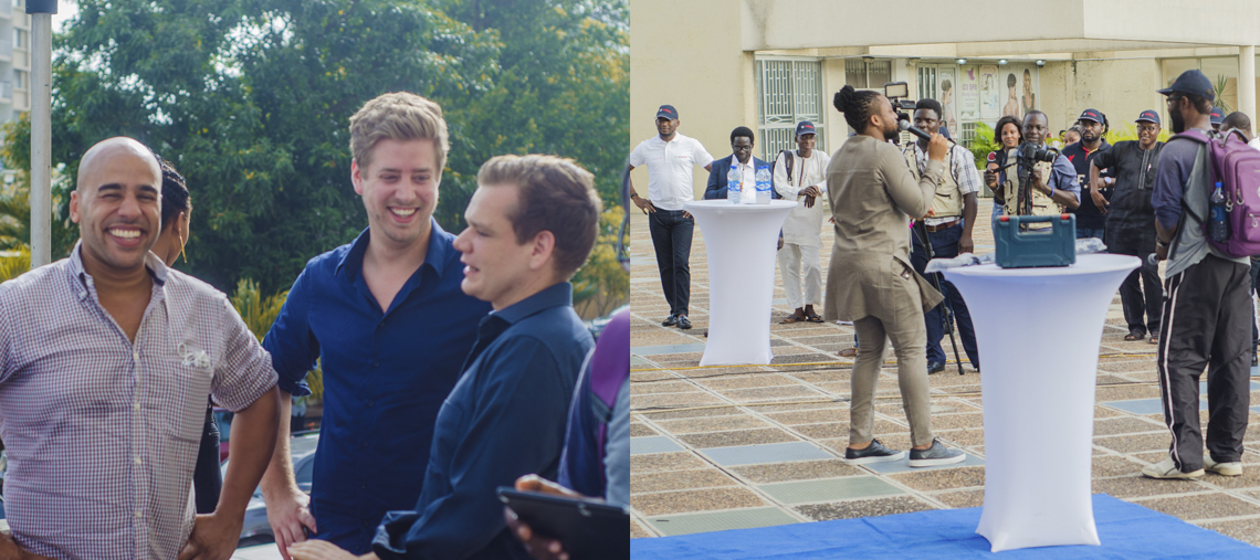 Two smiling men in casual clothes stand near a crowd at an outdoor event.