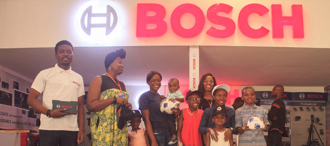 A group of adults and children pose with a Bosch sign in the background, smiling together.