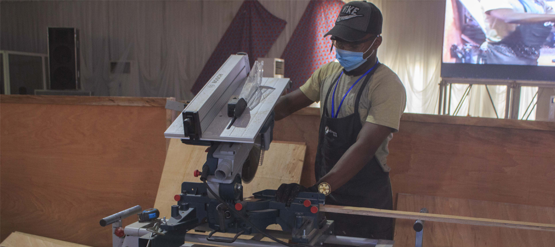 A man in a cap and mask operates a Bosch miter saw in a workshop with wooden panels.