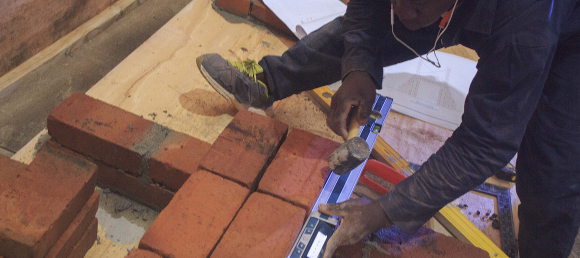 A person levels red bricks on a wooden floor using a hammer and a spirit level.