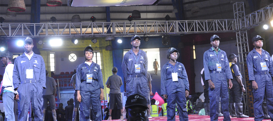 Six individuals in blue uniforms and caps stand on stage, under bright lights.