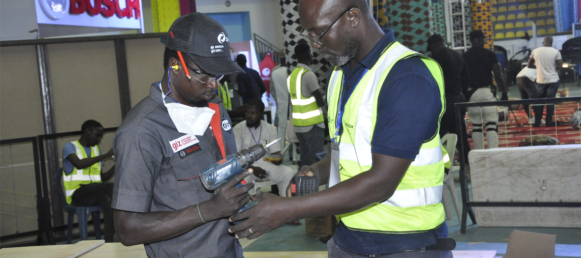 Two men inspect a Bosch tool at a workshop with safety vests and a colorful backdrop.