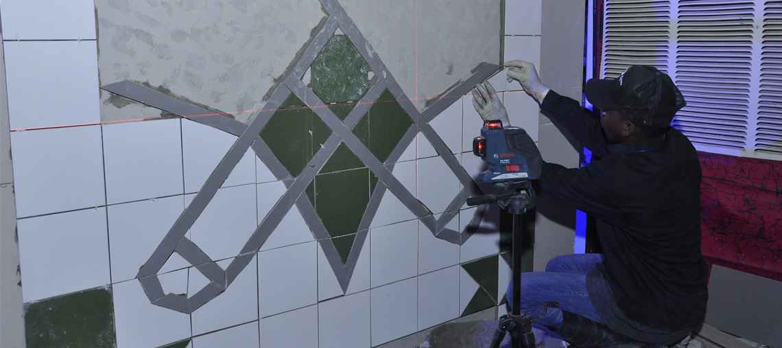 A worker uses a laser level on a tiled wall with green and white tiles.