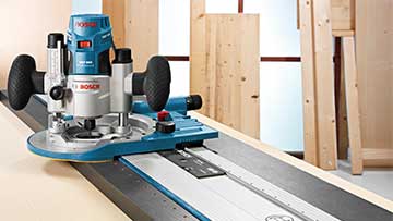 Een blauwe Bosch Professional bovenfrees staat op een tafel met houten planken op de achtergrond.