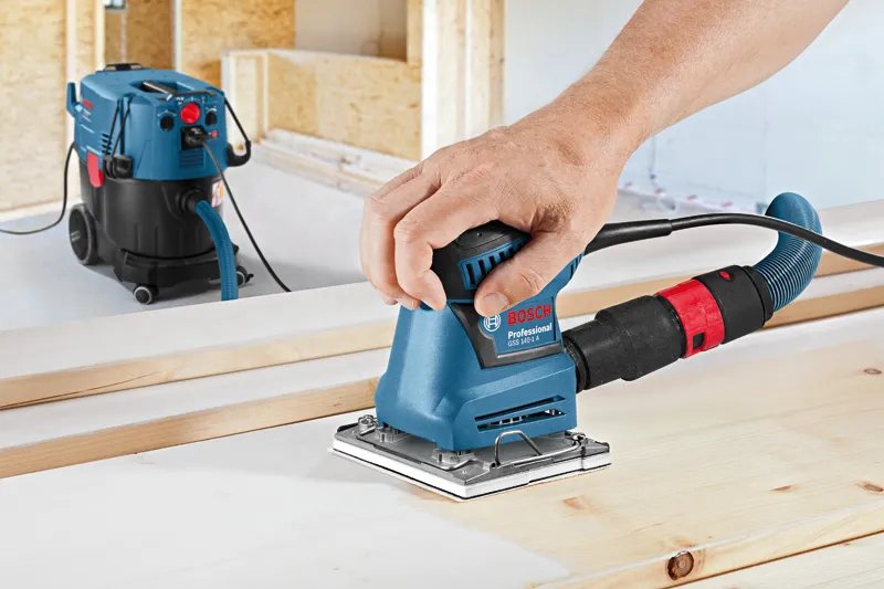 Iemand schuurt een houten plank met een vlakschuurmachine die is aangesloten op een stofafzuiging.