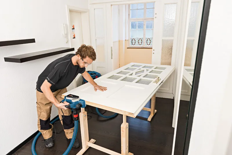 Een persoon die veiligheidsuitrusting draagt, gebruikt een schaafmachine op een witte houten deur.