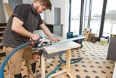 Een persoon met veiligheidsuitrusting zaagt een houten paneel met een accucirkelzaag op een werkbank.