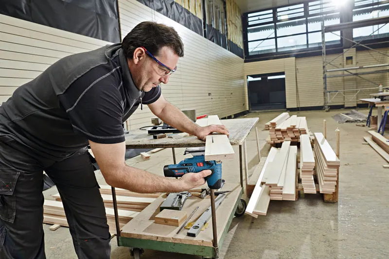 Een persoon met veiligheidsuitrusting zaagt hout met een accu-decoupeerzaag in een werkplaats.