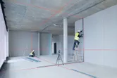 Twee personen met veiligheidsuitrusting installeren gipsplaten in een kamer met behulp van een laserwaterpas.