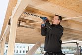 Een persoon met veiligheidsuitrusting boort in een houten balk op een bouwplaats.