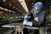Een persoon met veiligheidsuitrusting boort in een metalen balk in een fabriek.