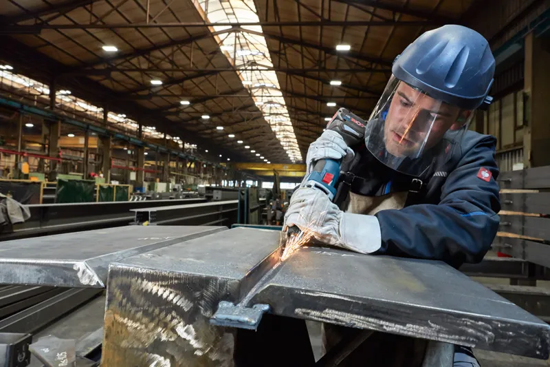 Een persoon die veiligheidsuitrusting draagt, slijpt metaal met een elektrisch gereedschap in een fabriek.