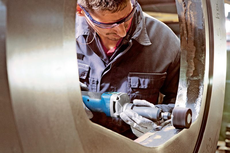 Een persoon met veiligheidsuitrusting polijst een grote metalen ring met een snoerloze rechte slijper.
