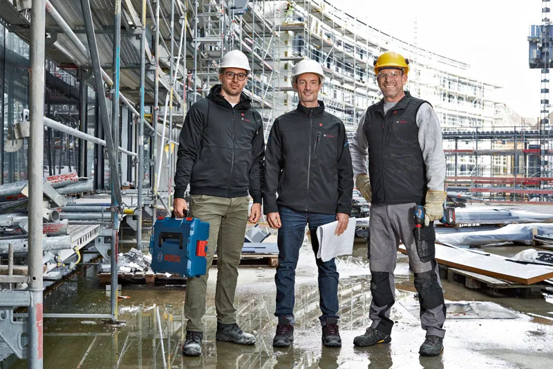Drie arbeiders met veiligheidsuitrusting staan op een bouwplaats, één van hen houdt een elektrisch gereedschap vast.