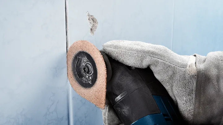 Persoon met veiligheidsuitrusting slijpt met roterend gereedschap een gat in een tegel.