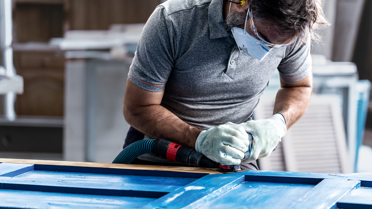 Een persoon met veiligheidsuitrusting schuurt een blauw houten oppervlak met een elektrisch gereedschap.