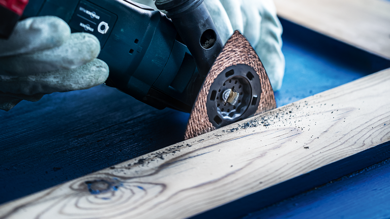 Een persoon met veiligheidsuitrusting schuurt een houten plank met een multitool.