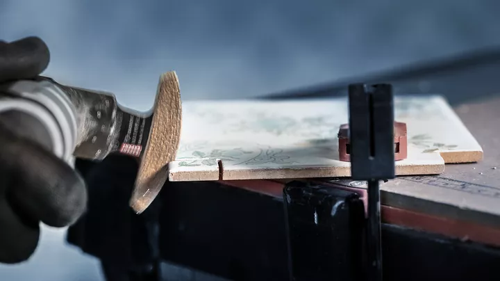 Een persoon met veiligheidsuitrusting snijdt een keramische tegel met behulp van een elektrisch oscillerend gereedschap.